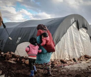 A woman and children walk near tents in a refugee camp in Idlib, showcasing resilience amidst adversity.