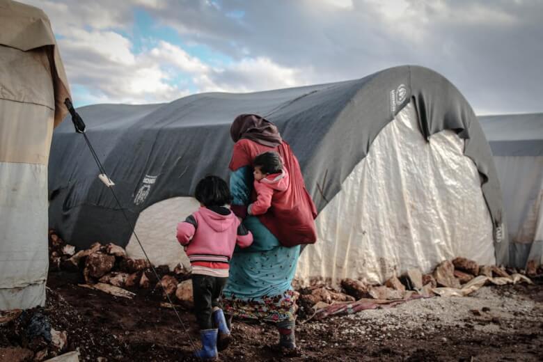 A woman and children walk near tents in a refugee camp in Idlib, showcasing resilience amidst adversity.