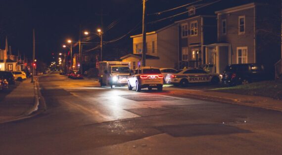 Cars with glowing lights parked on asphalt road near police car placed near residential building in small town in evening