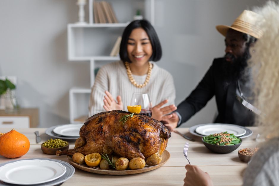 Cheerful multiethnic people having dinner together at table with roasted turkey while celebrating Thanksgiving Day
