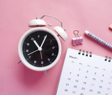Flat lay of a pink-themed workspace featuring an alarm clock, calendar, pen, and paper clip on a pastel surface.