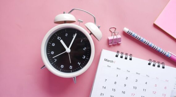 Flat lay of a pink-themed workspace featuring an alarm clock, calendar, pen, and paper clip on a pastel surface.
